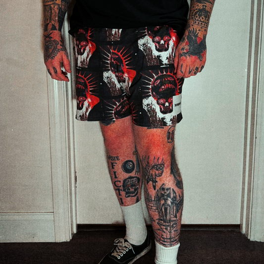A person wearing black graphic shorts with a red, white, and black skull-and-sunburst print, paired with a black t-shirt, white socks, and black sneakers, standing in front of a white door.